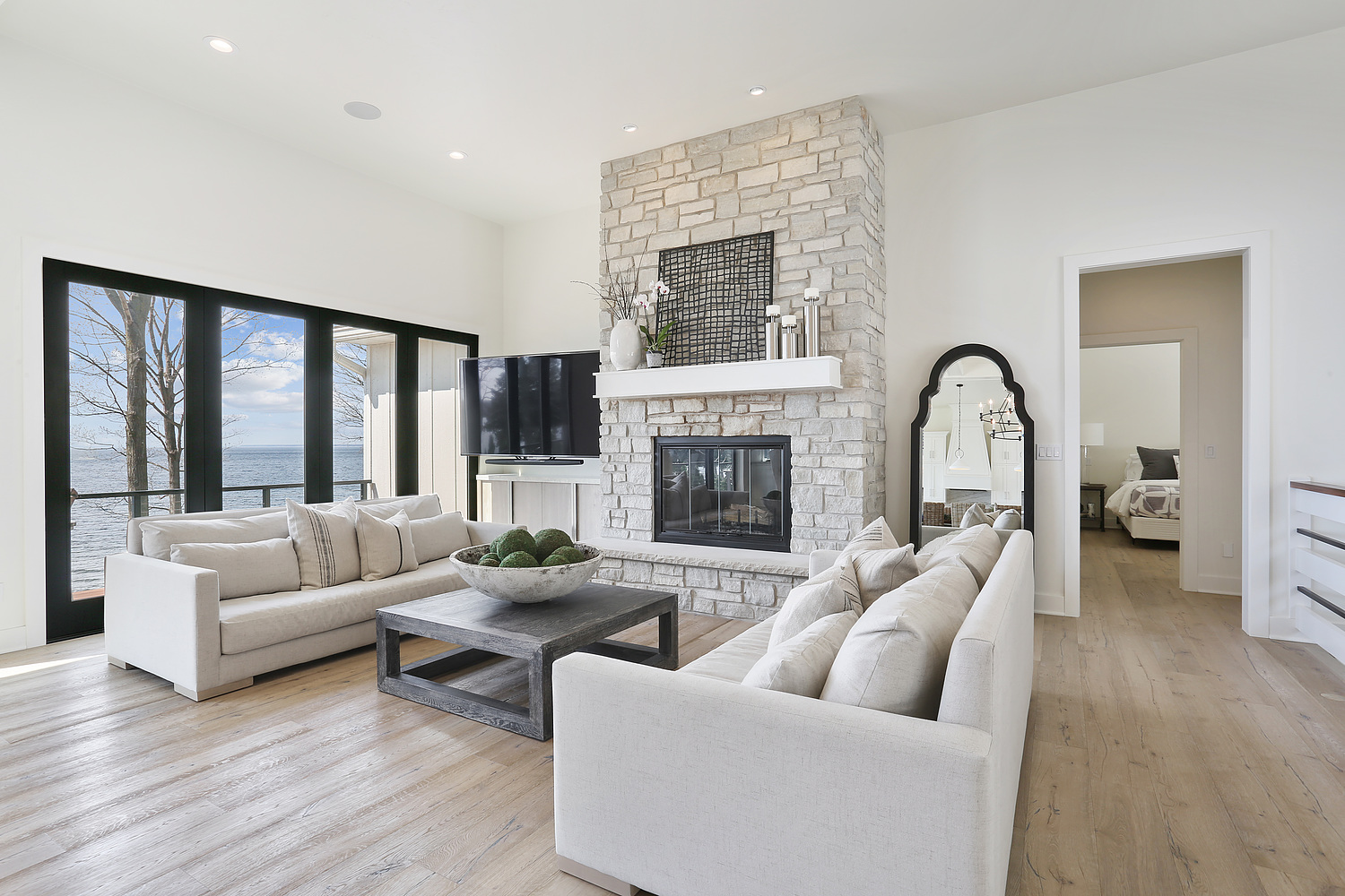 A neutral colored living room with grey stone fireplace in centered on wall, windows overlooking water