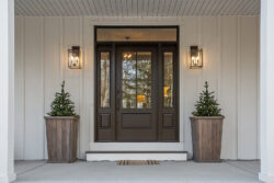 A dark brown front door with a window, two shrubs in planters on either side and amber porch lights