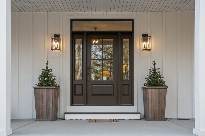 A dark brown front door with a window, two shrubs in planters on either side and amber porch lights
