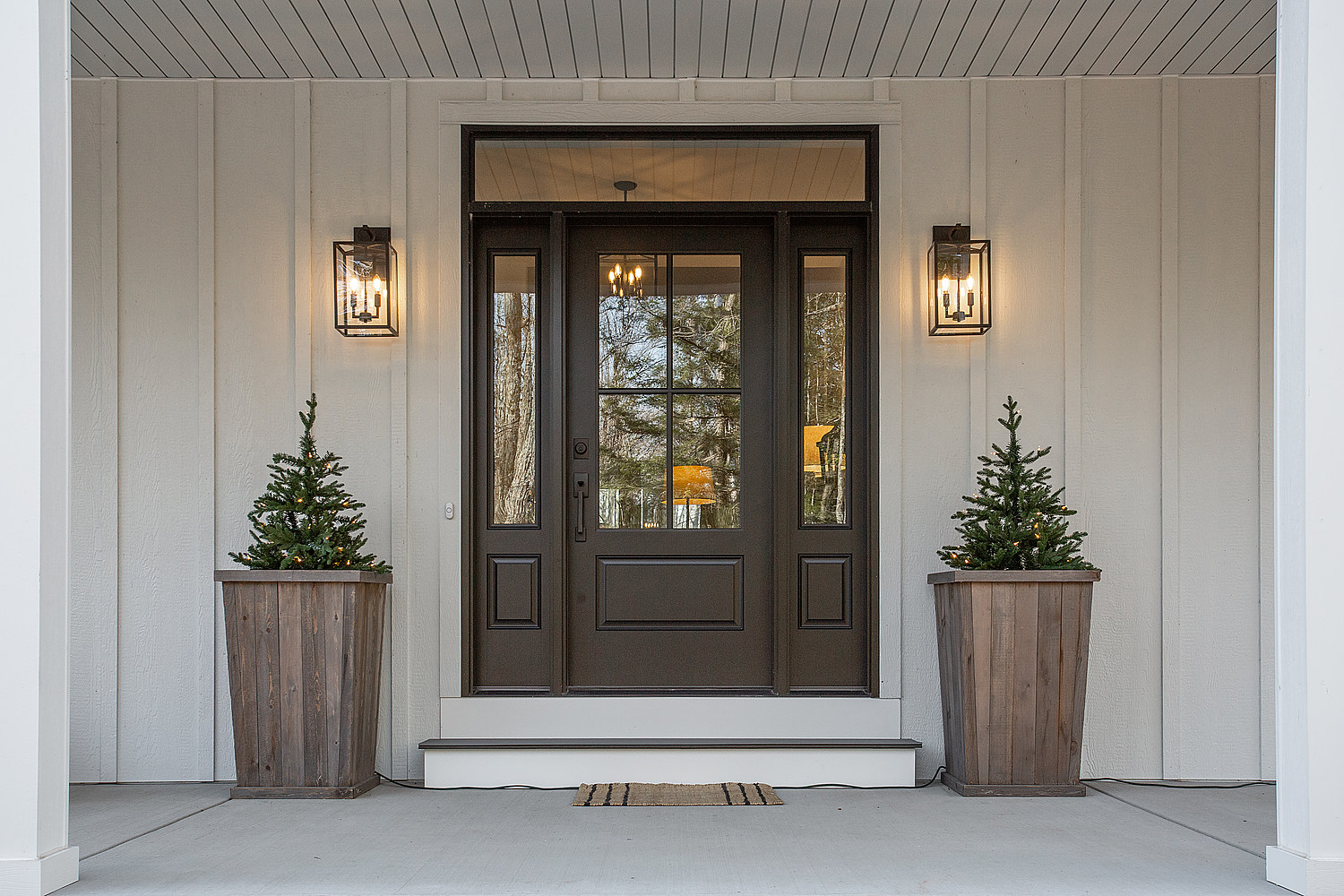 A dark brown front door with a window, two shrubs in planters on either side and amber porch lights